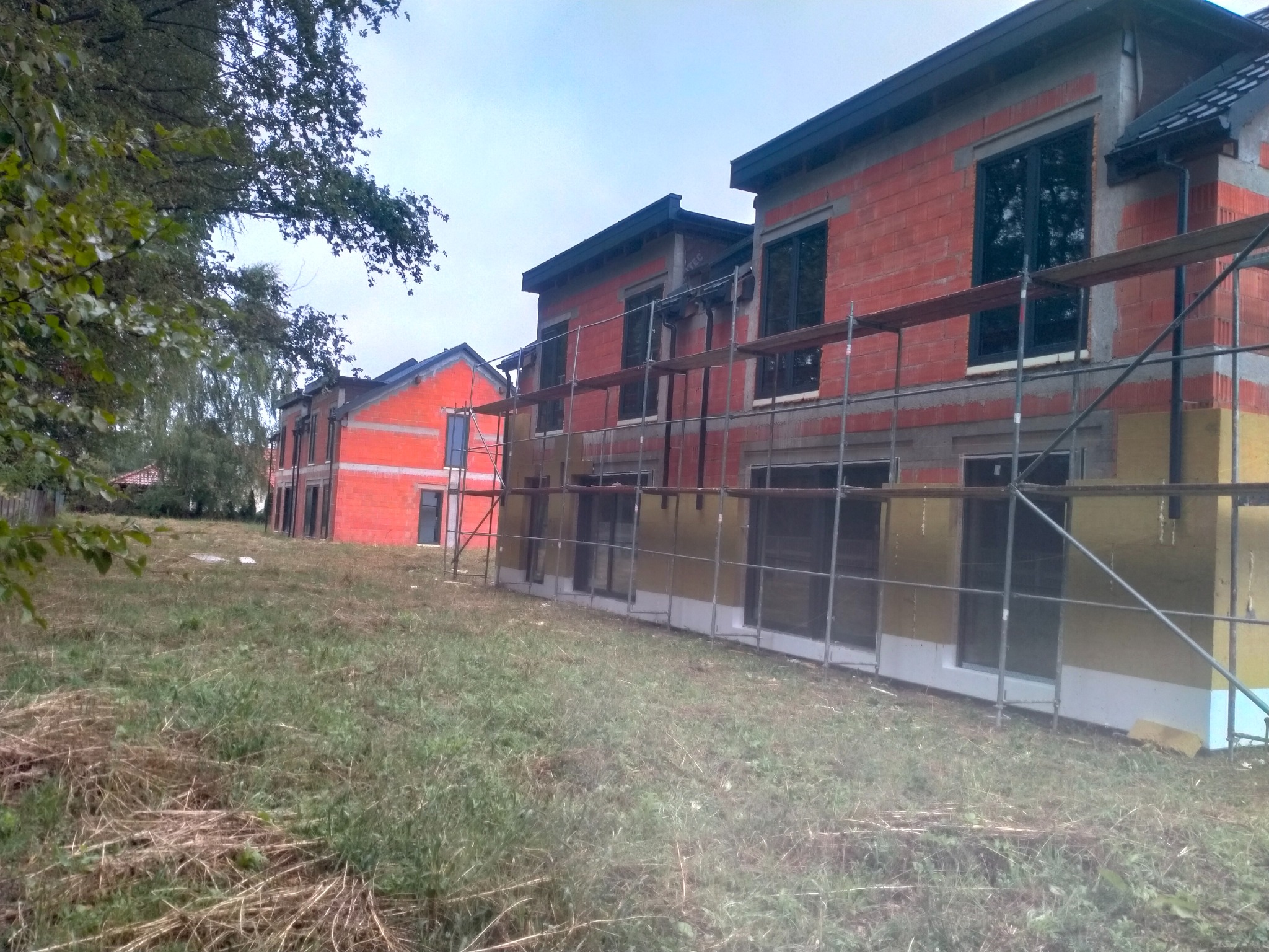 Budowa szeregowców z cegły, widoczne rusztowania i ocieplenie fundamentów. Surowy stan, teren zarośnięty trawą. Ujęcie z dołu, pochmurne niebo.