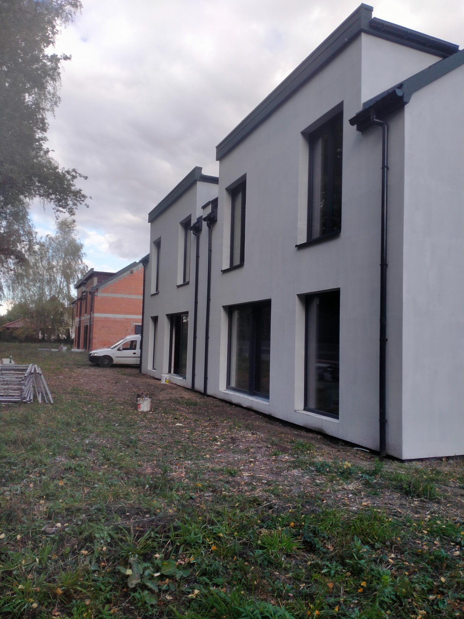 Nowoczesny, dwukondygnacyjny dom z szarą elewacją i ciemnymi oknami. W tle budynek w stanie surowym i biały samochód dostawczy. Widoczne rusztowania i puszka farby.