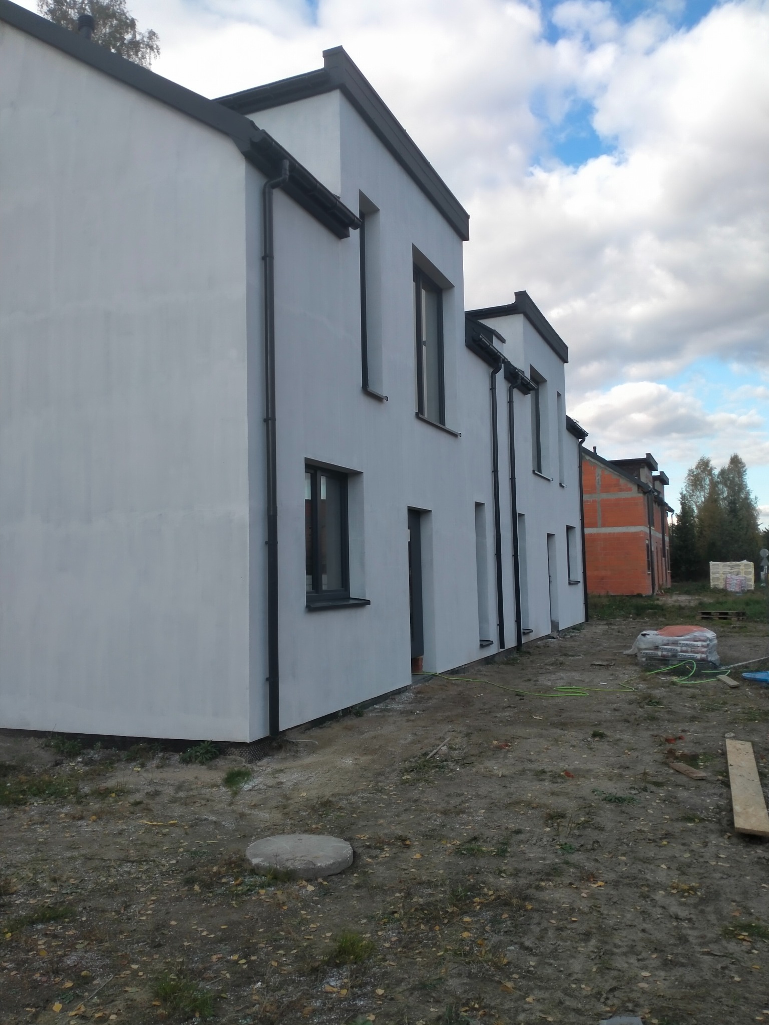 Biała elewacja szeregowca z czarnymi rynnami i oknami, widok z boku na tle błękitnego nieba z chmurami. W tle budynek w trakcie budowy z czerwonej cegły.