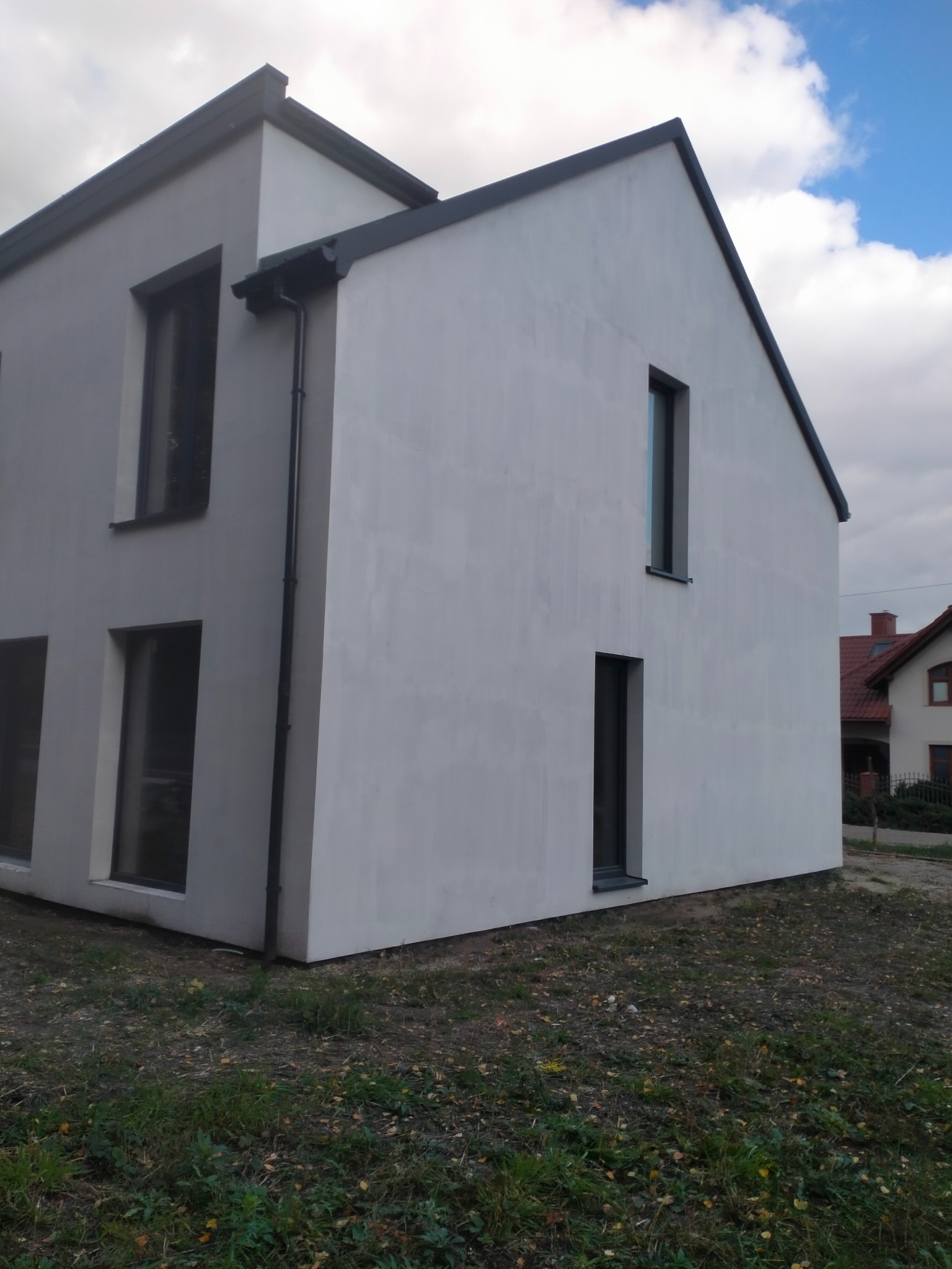 Nowoczesny dom jednorodzinny z jasną elewacją i ciemnymi ramami okiennymi, widok z zewnątrz, trawa z opadłymi liśćmi u podstawy budynku.