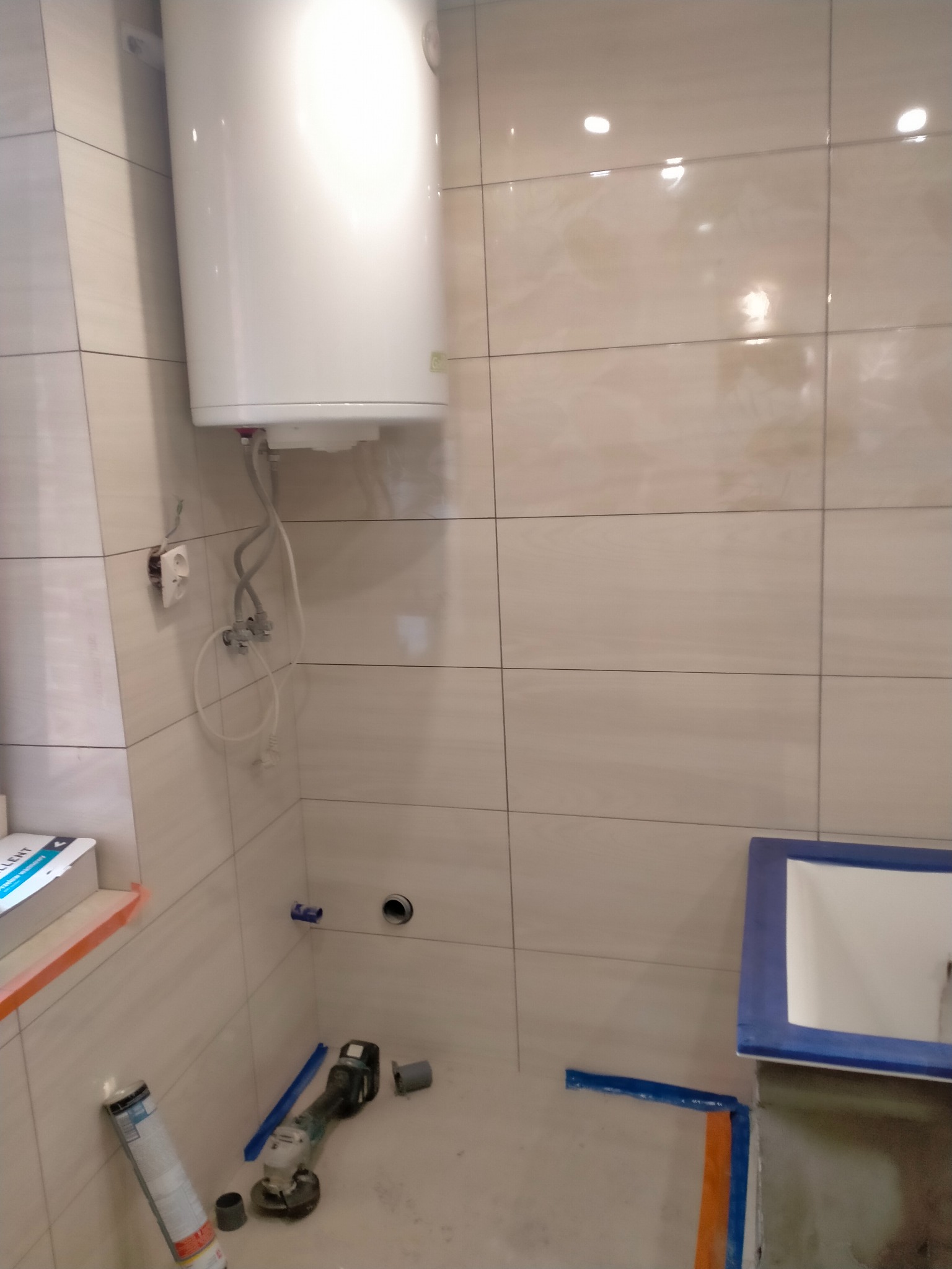 Biały bojler zawieszony na ścianie z beżowych płytek w łazience w trakcie remontu. Widoczne narzędzia i niedokończona instalacja umywalki.