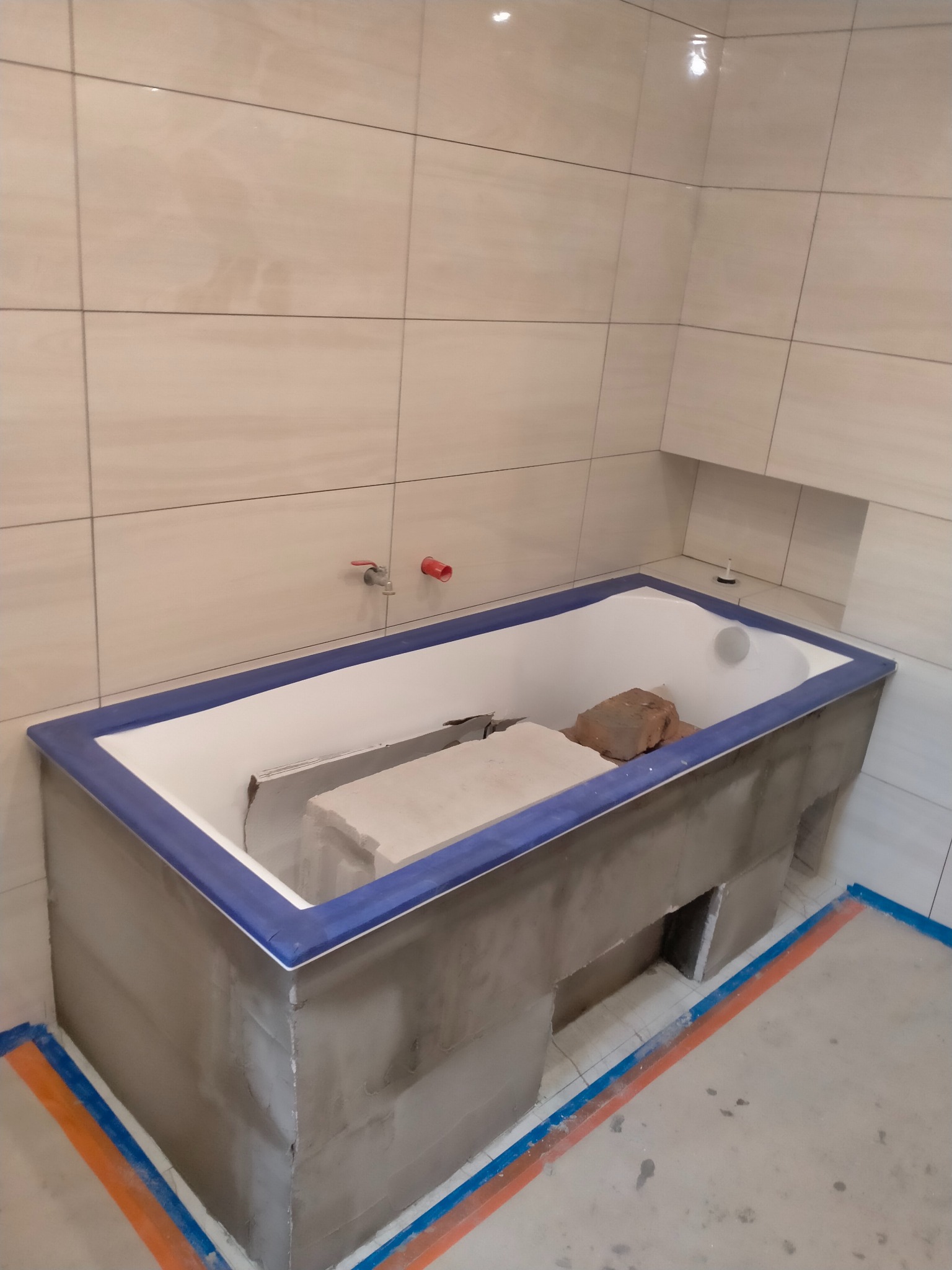 Wanna obudowana szarym betonem i wykończona niebieską listwą w łazience z beżowymi płytkami. Prace glazurnicze w Rękoraju na etapie obudowy wanny.