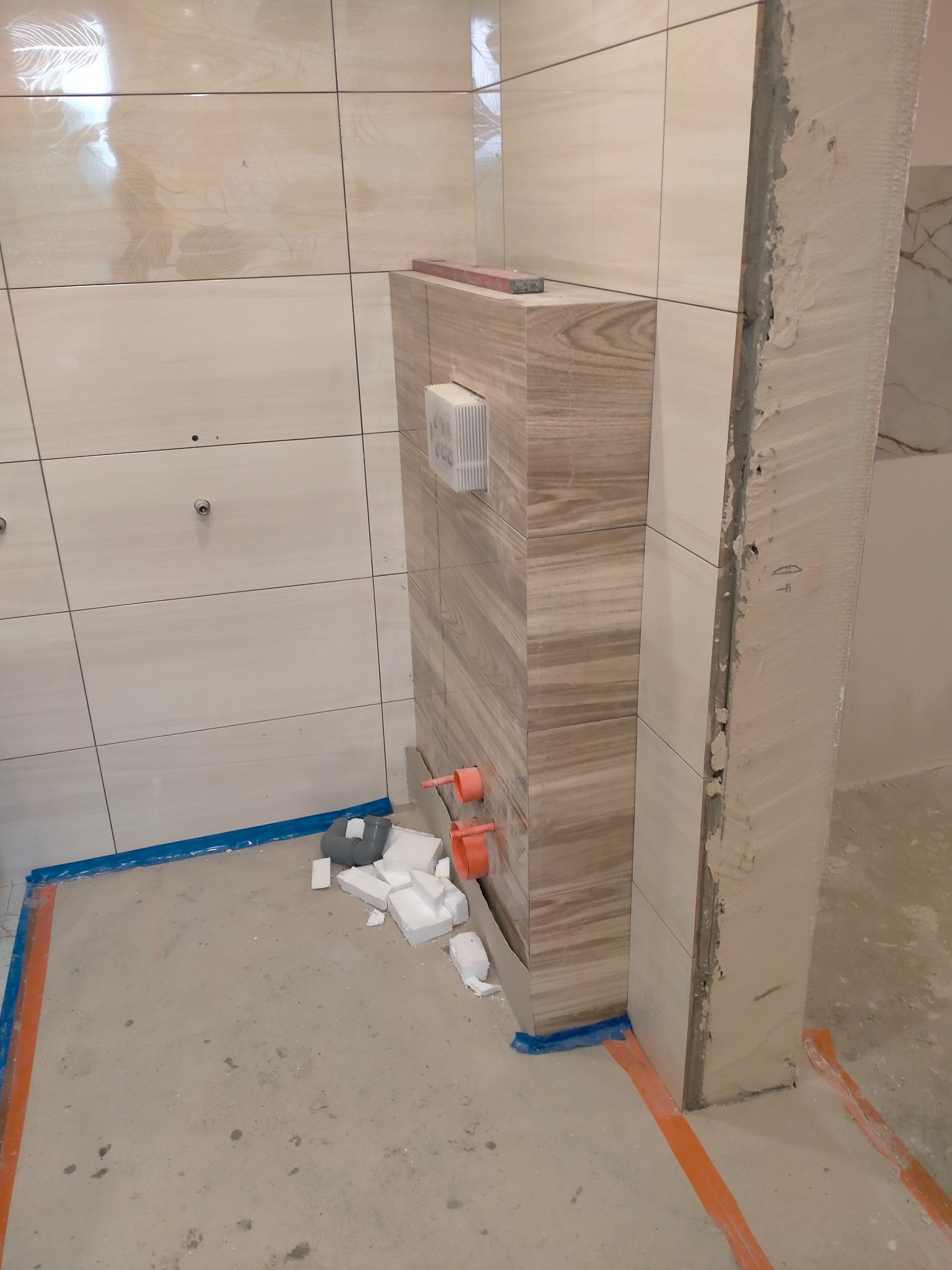 Wykończenie łazienki: beżowe płytki imitujące marmur na ścianach i obudowie stelaża WC. Widoczne fragmenty instalacji wodnej i przygotowane podłoże pod posadzkę.