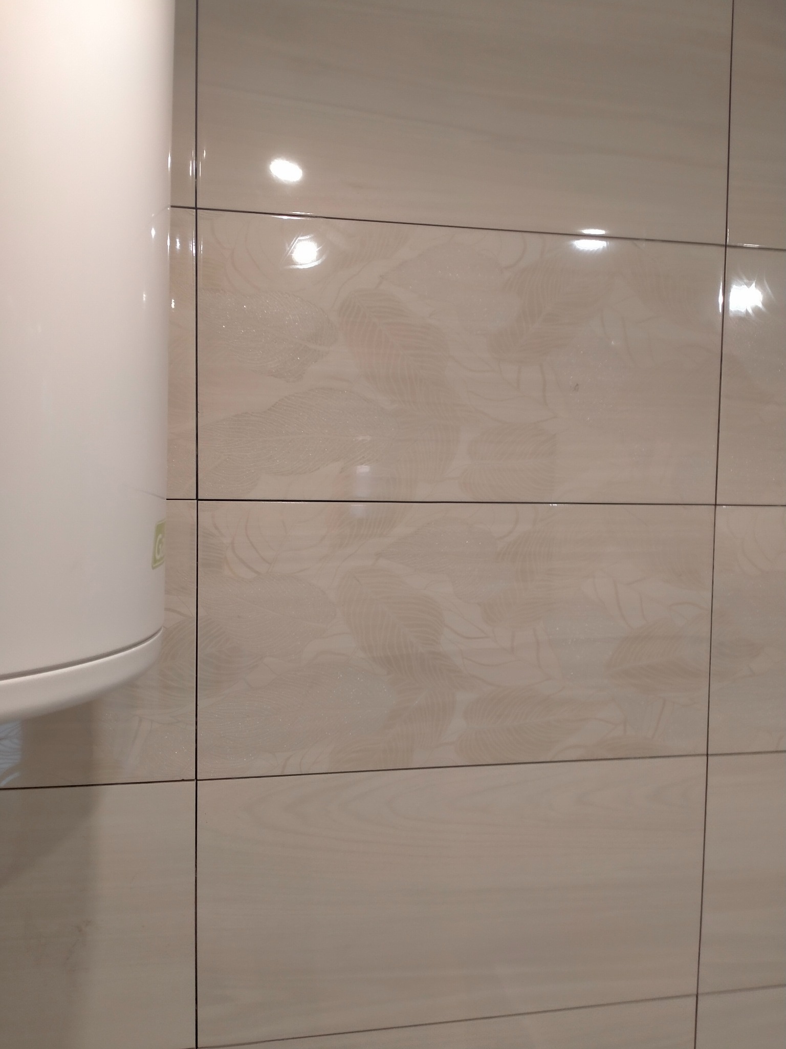 Beżowe, błyszczące płytki ścienne z delikatnym wzorem liści, ułożone poziomo. Fragment bojlera widoczny po lewej stronie. Oświetlenie odbija się w powierzchni płytek.