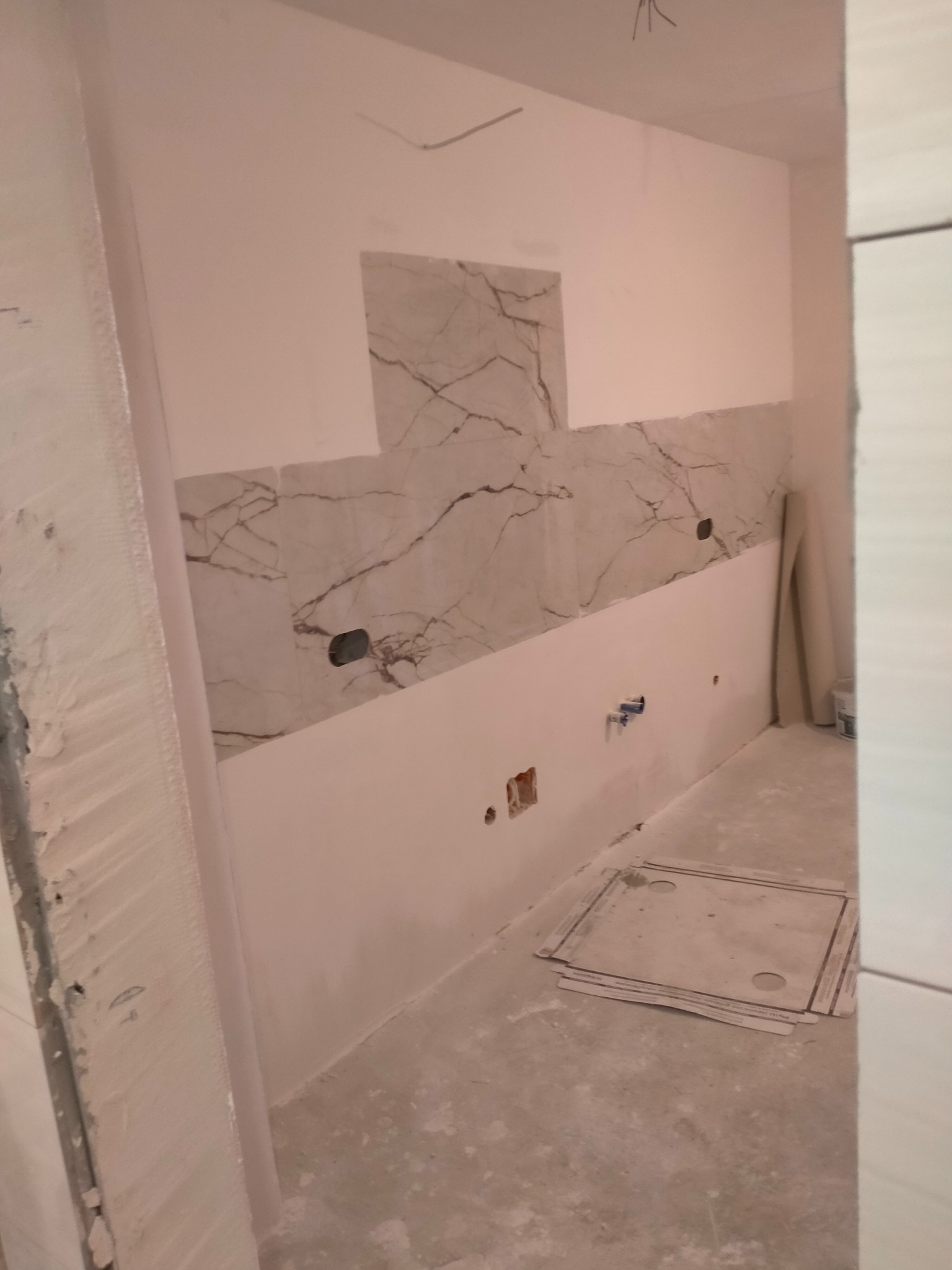 Fragment ściany z częściowo położonymi płytkami imitującymi marmur, widoczne otwory na gniazdka i rury, podłoga w trakcie prac wykończeniowych.