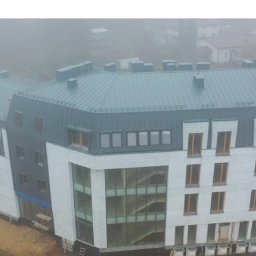 Nowoczesny budynek wielorodzinny z szarą blachą na dachu, widoczny w mglisty dzień. Częściowo widoczne rusztowania i teren budowy.