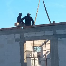 Budowa domu z betonu komórkowego: dwóch robotników na ścianie, dźwig podnosi element konstrukcyjny. Widoczne okno z metalową ramą i niebieskie niebo.