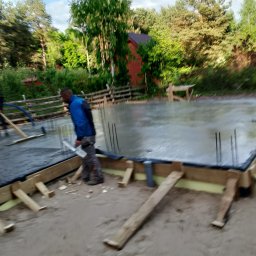 Wylewka fundamentów pod dom jednorodzinny w Warszawie. Widoczne zbrojenia i szalunki. Prace budowlane w toku, teren zalesiony w tle.