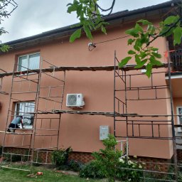 Ocieplony dom w kolorze terakoty z rusztowaniem. Pracownik na rusztowaniu przy oknie. Widoczny klimatyzator na elewacji. Fragment balkonu z ozdobną balustradą.