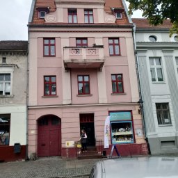 Nelav Sp Zo.o. - Odnowiona, różowa kamienica z czerwonymi oknami i balkonem, sąsiadująca z budynkami w różnym stylu. Widoczny fragment chodnika i zaparkowany samochód.