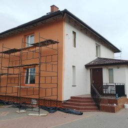 Nelav Sp Zo.o. - Dwukondygnacyjny dom w trakcie malowania elewacji. Część budynku w kolorze pomarańczowym, druga część biała. Widoczne rusztowania i schody wejściowe.
