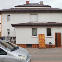 Nelav Sp Zo.o. - Dwupiętrowy dom z brązowym dachem w trakcie remontu elewacji, widoczne rusztowania i stara drewniana brama oparta o ścianę. Na pierwszym planie srebrny samochód.