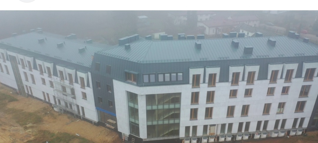 Nowoczesny budynek wielorodzinny z szarą blachą na dachu, widoczny w mglisty dzień. Częściowo widoczne rusztowania i teren budowy.