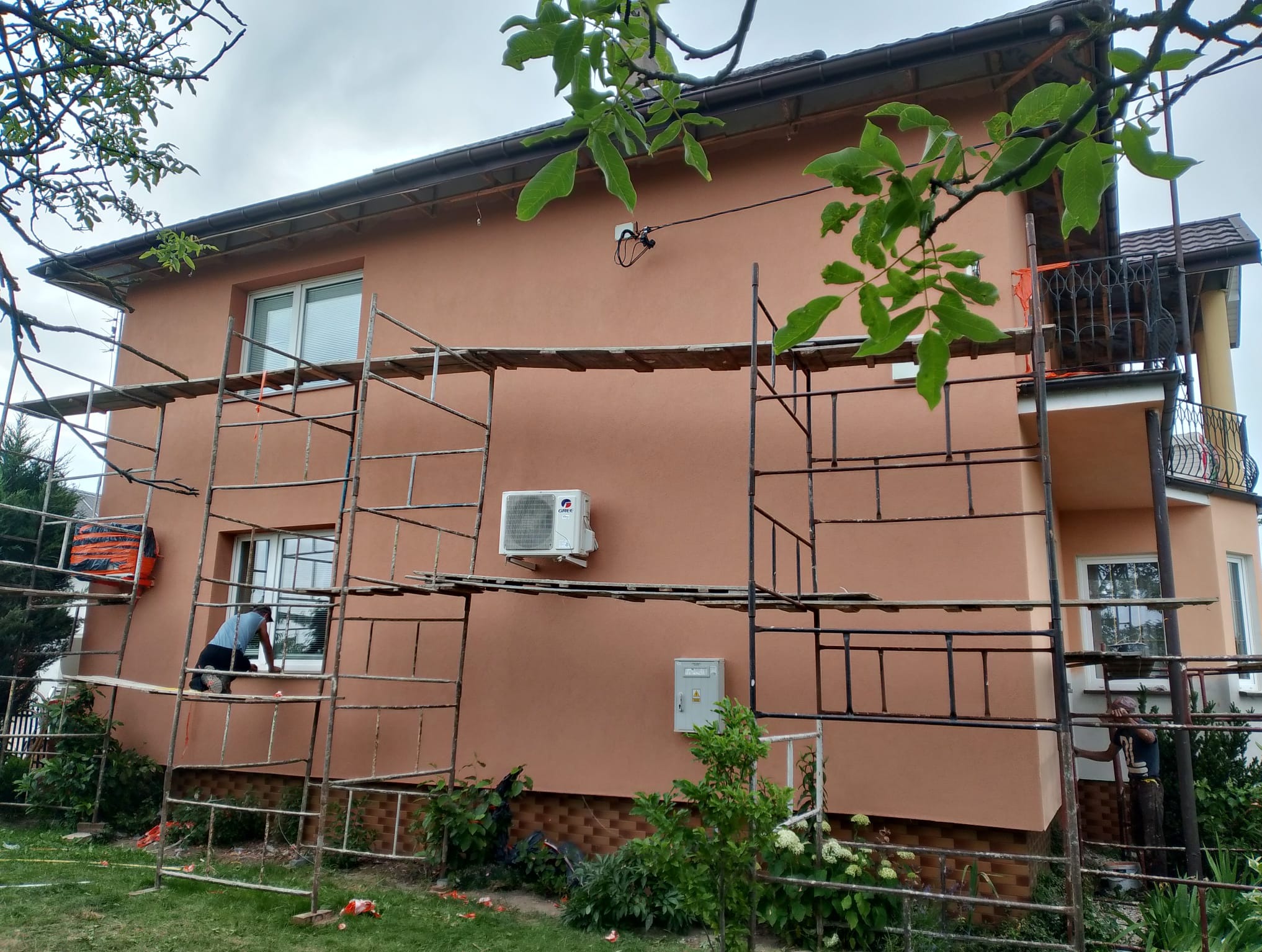 Ocieplony dom w kolorze terakoty z rusztowaniem. Pracownik na rusztowaniu przy oknie. Widoczny klimatyzator na elewacji. Fragment balkonu z ozdobną balustradą.