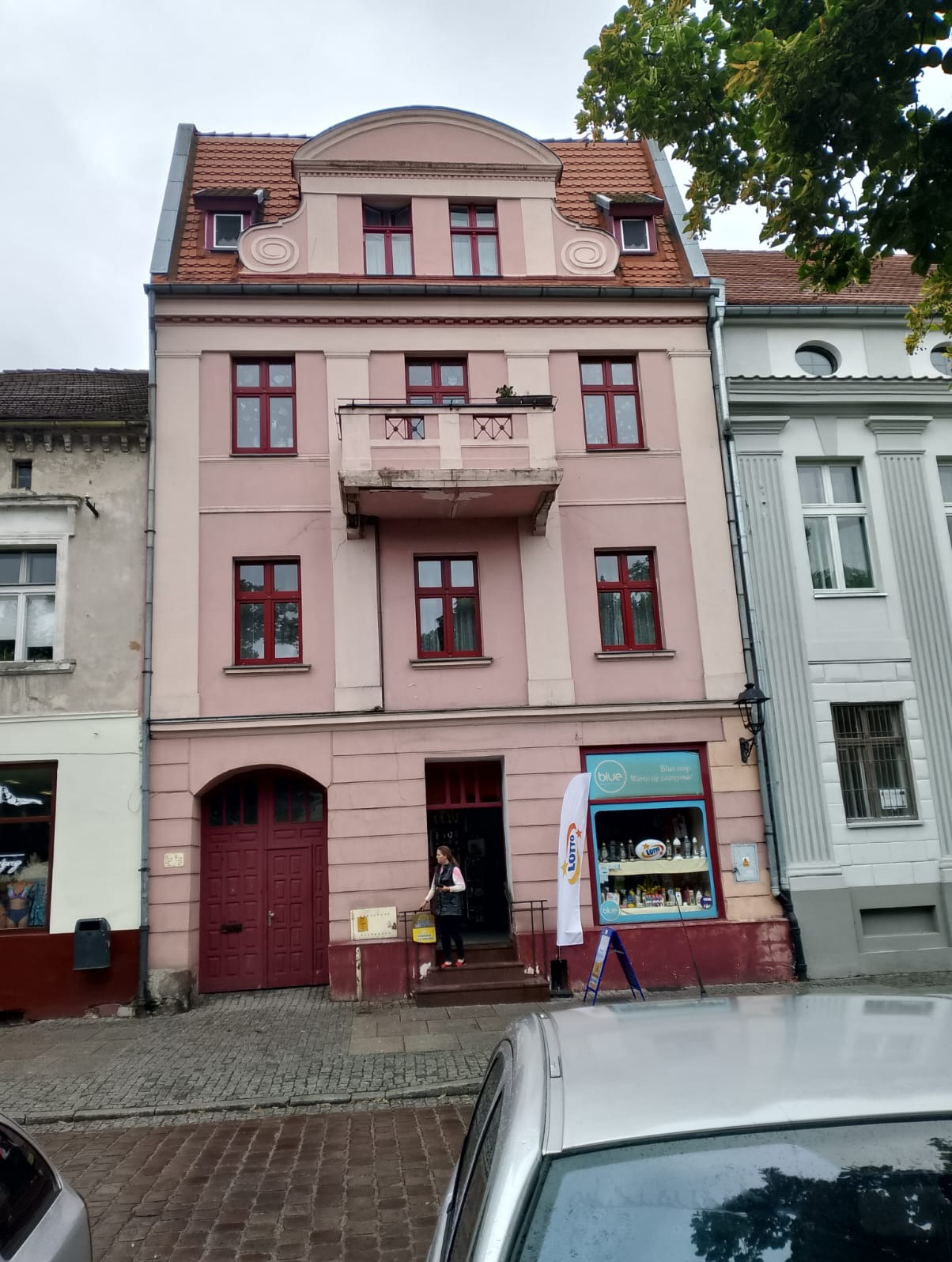 Odnowiona, różowa kamienica z czerwonymi oknami i balkonem, sąsiadująca z budynkami w różnym stylu. Widoczny fragment chodnika i zaparkowany samochód.