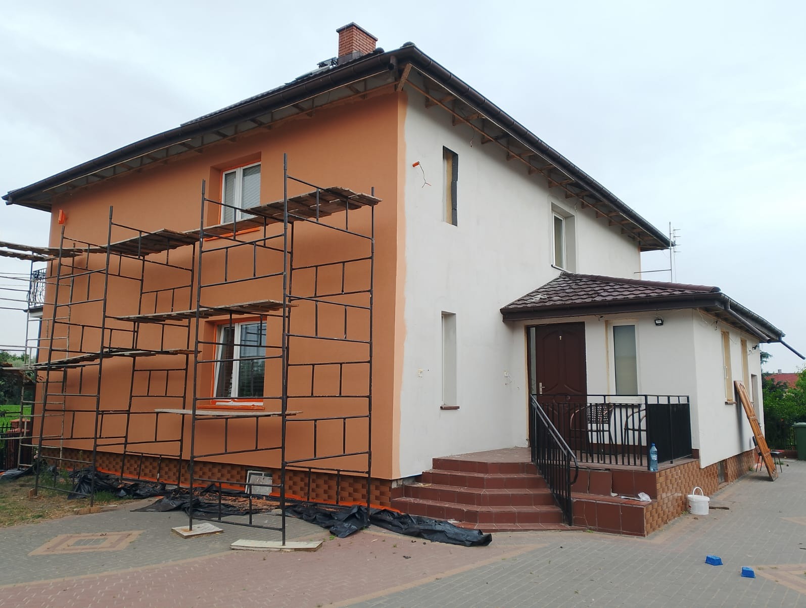 Dwukondygnacyjny dom w trakcie malowania elewacji. Część budynku w kolorze pomarańczowym, druga część biała. Widoczne rusztowania i schody wejściowe.