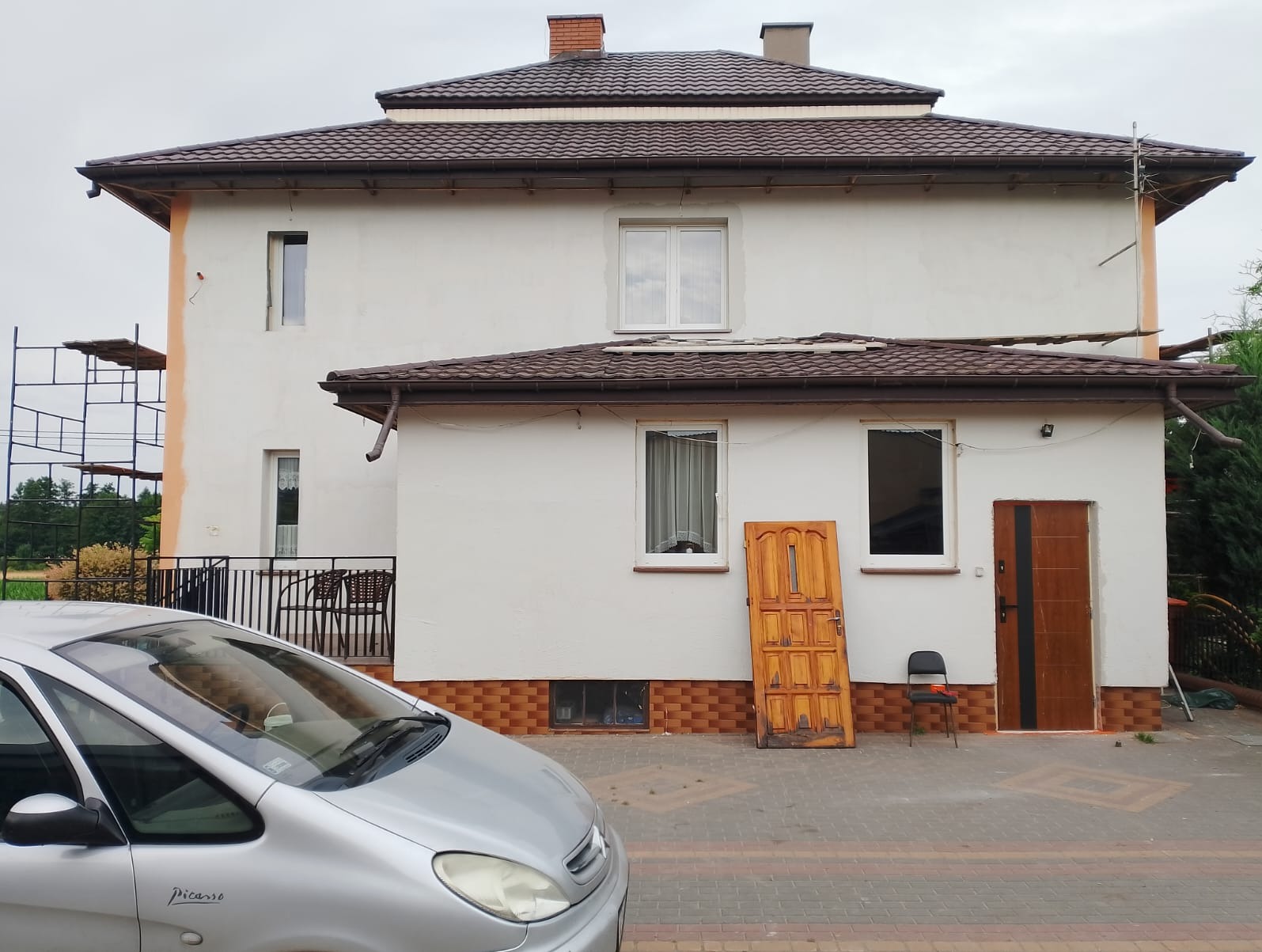 Dwupiętrowy dom z brązowym dachem w trakcie remontu elewacji, widoczne rusztowania i stara drewniana brama oparta o ścianę. Na pierwszym planie srebrny samochód.