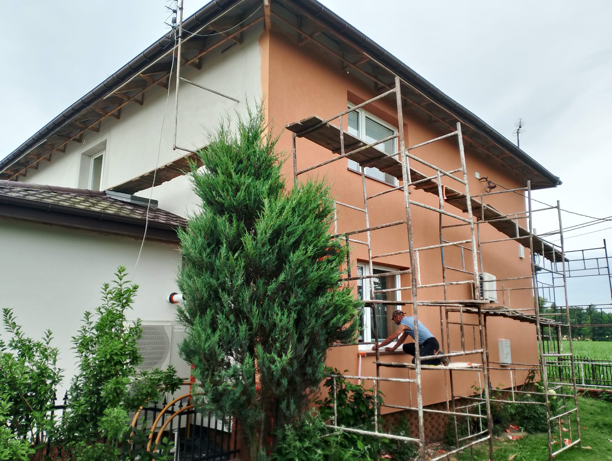 Malowanie elewacji domu jednorodzinnego z rusztowania. Widoczny fragment pomalowanej ściany na biało i pracownik przy oknie. W tle zieleń i błękitne niebo.