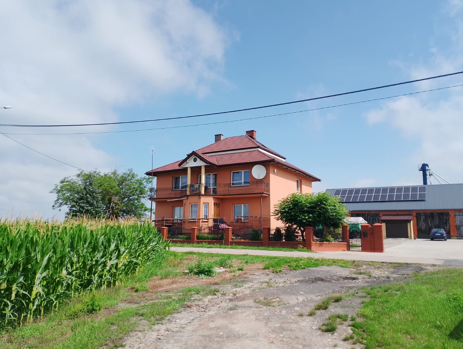 Dwukondygnacyjny dom z balkonami, pomalowany na brzoskwiniowo, z czerwoną dachówką. Na dachu antena satelitarna. Obok budynek gospodarczy z panelami słonecznymi.
