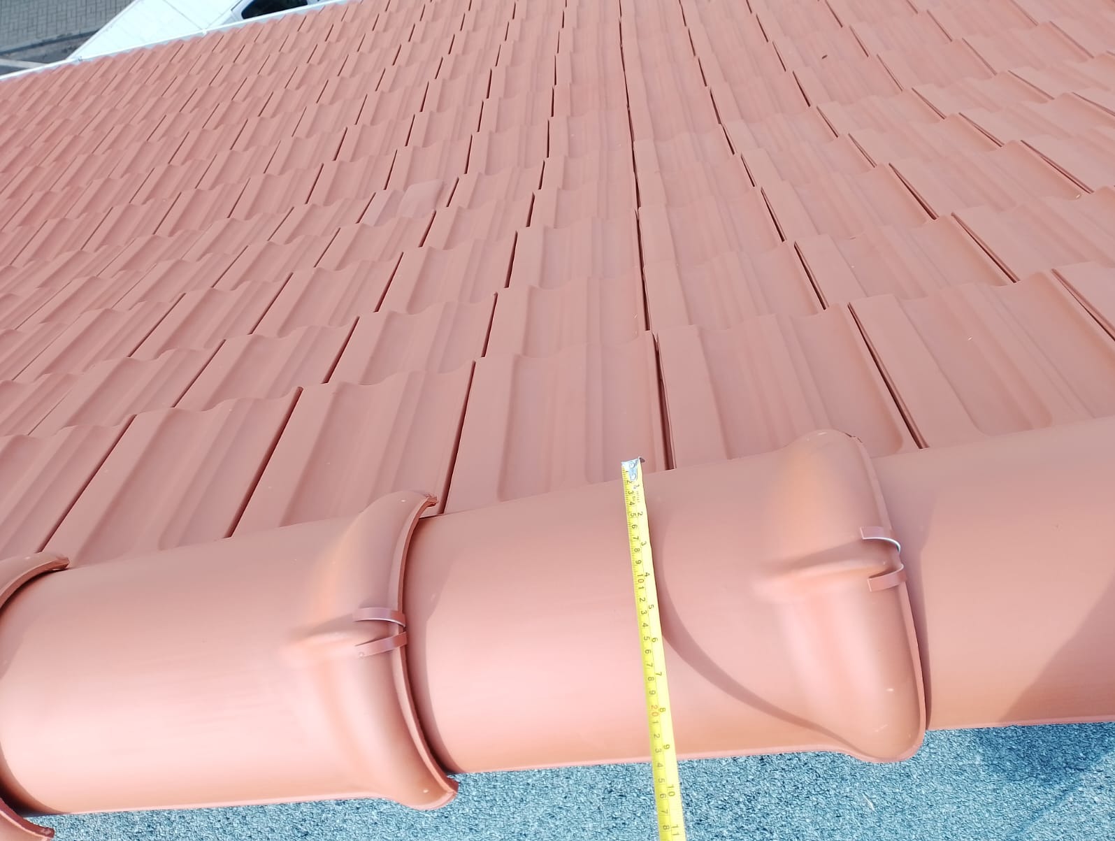 Czerwona dachówka ceramiczna z bliska, z widoczną rynną i rozłożoną miarką. Układanie dachówki na dachu, pomiar odległości.