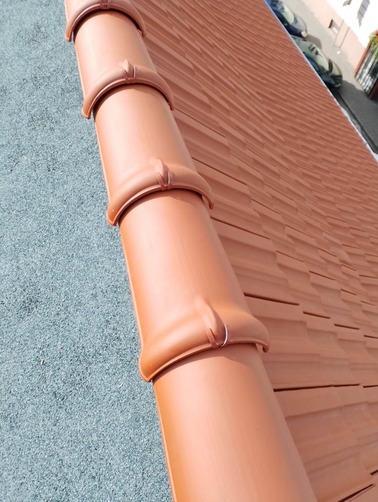 Ceglasty dach z ceramicznej dachówki karpiówki i rynna z obejmami, widok z góry, lekki skos, szara podsypka, fragment ulicy w tle.