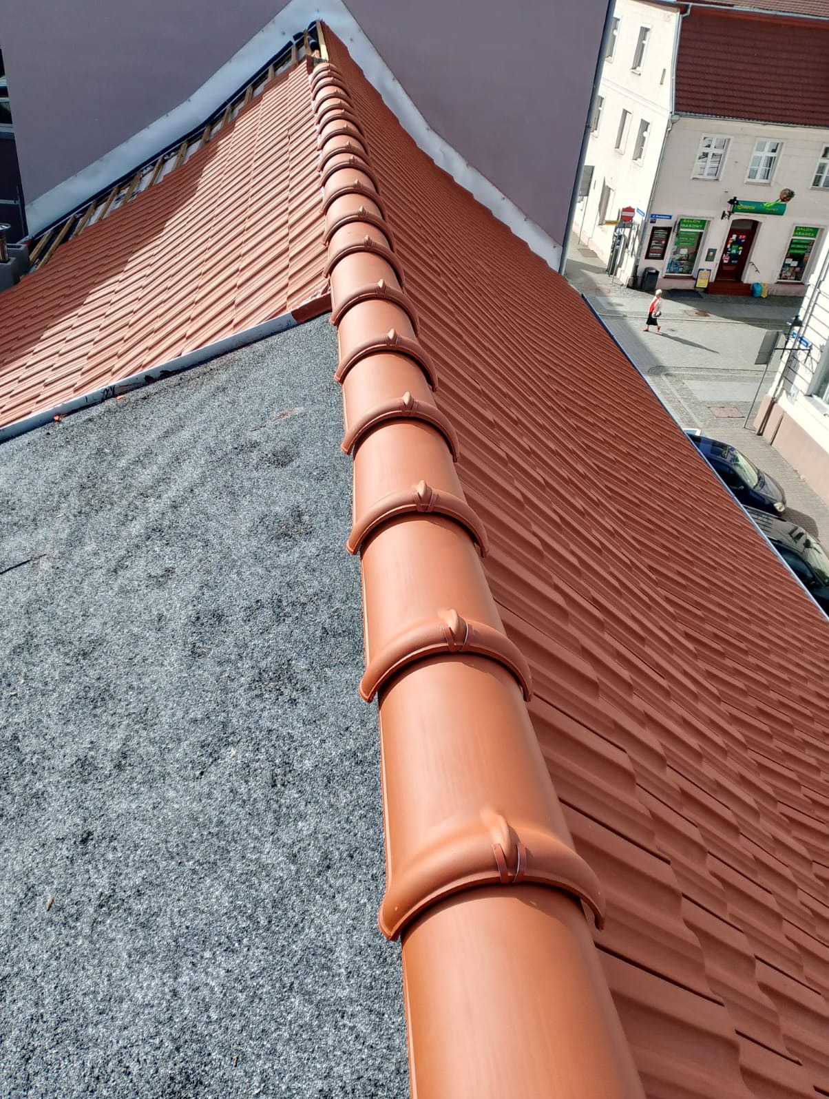 Czerwona dachówka ceramiczna na dachu budynku, widok z góry, z elementami obróbki blacharskiej i fragmentem ulicy w tle. Układana dachówka w trakcie prac dekarskich.