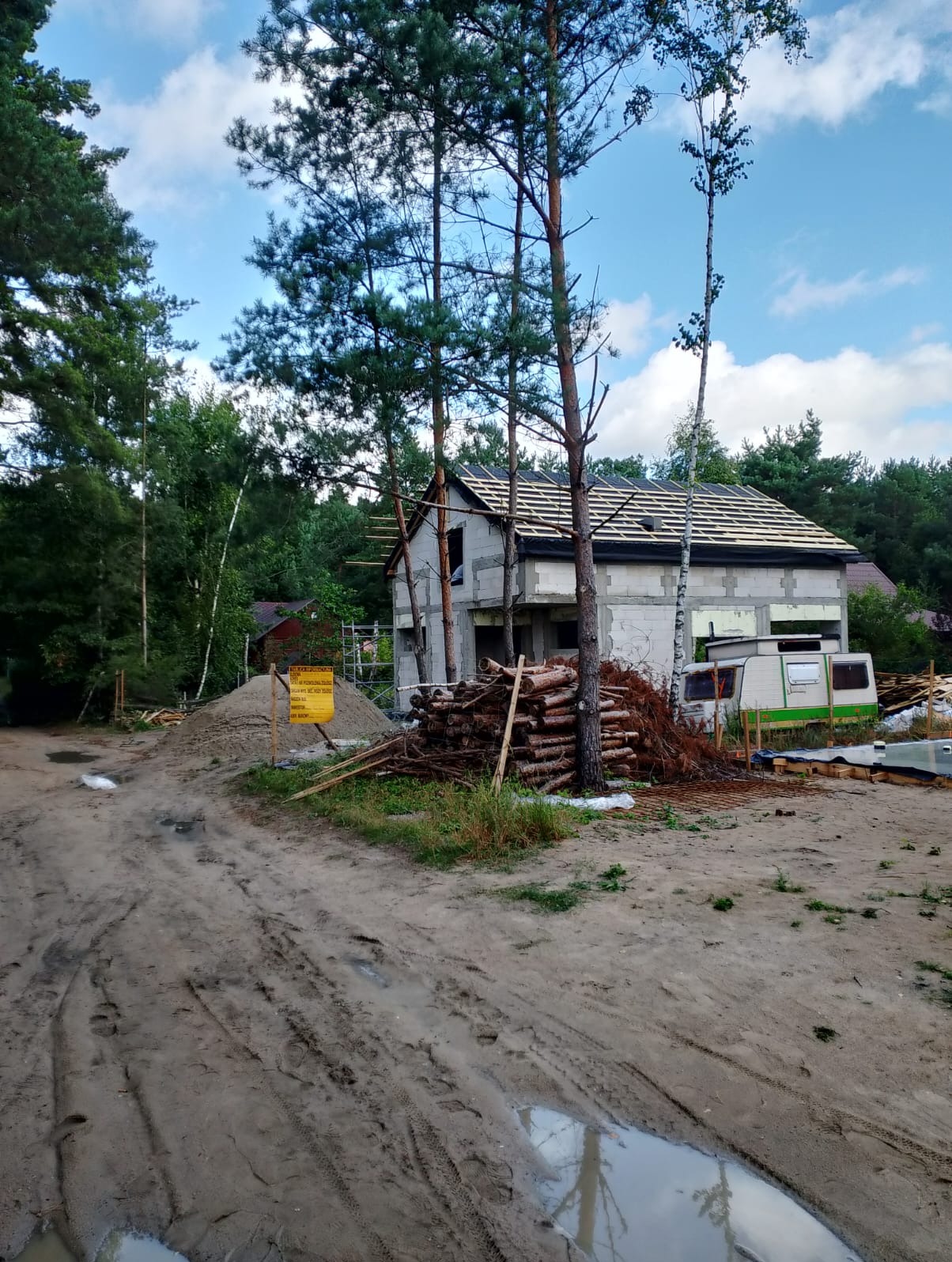 Surowy dom murowany w trakcie budowy, z widocznym drewnianym szkieletem dachu i stertą drewna na posesji, otoczony zielenią drzew. W tle przyczepa kempingowa.