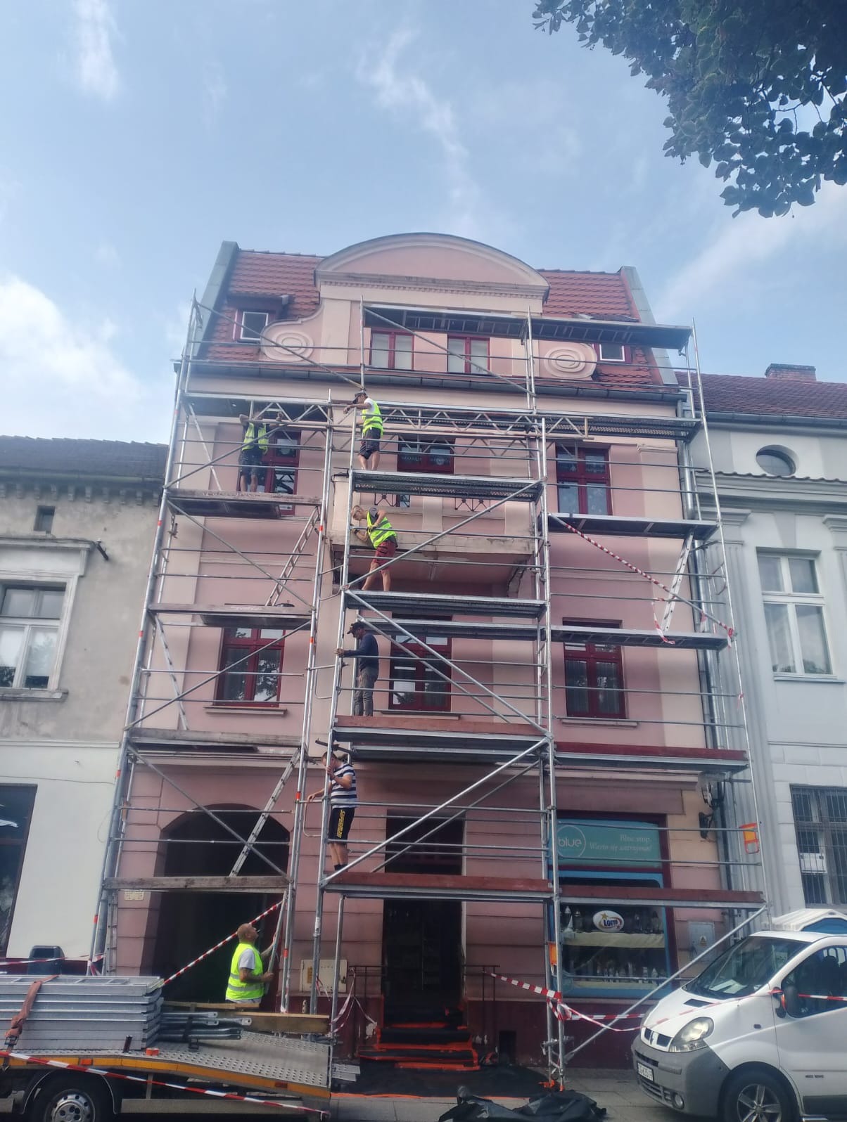 Praca na rusztowaniu przy odnowie elewacji trzypiętrowego budynku z czerwonym dachem. Pracownicy w kamizelkach. Na pierwszym planie przyczepa i samochód dostawczy.
