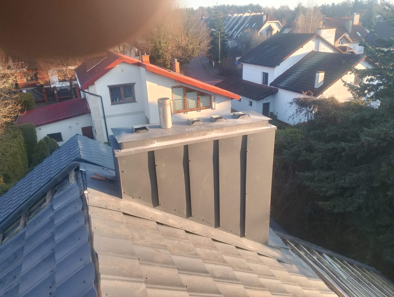 Szary komin z blachy na dachu z dachówki bitumicznej, w tle zabudowa jednorodzinna w słoneczny dzień. Widok z góry.