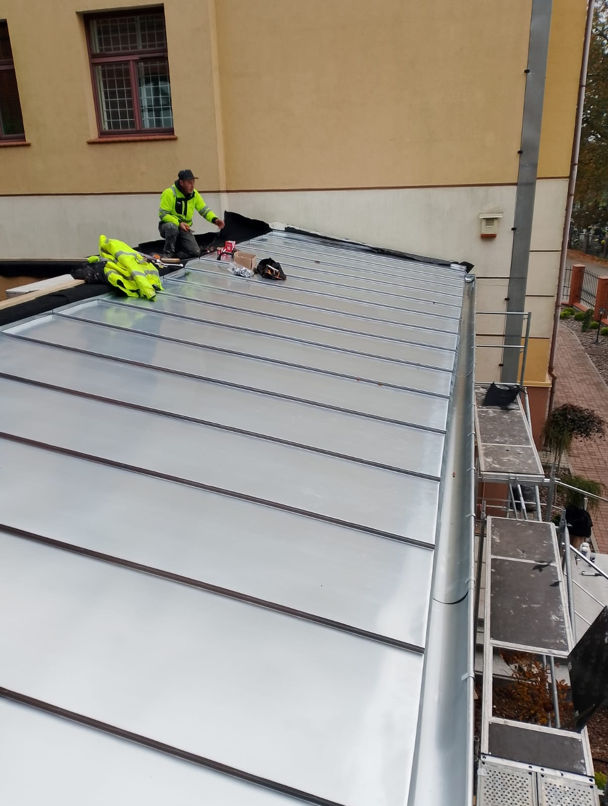 Montaż blachy na dachu budynku. Pracownik w odblaskowej kurtce układa panele. Widoczne narzędzia, rusztowanie i fragment elewacji.