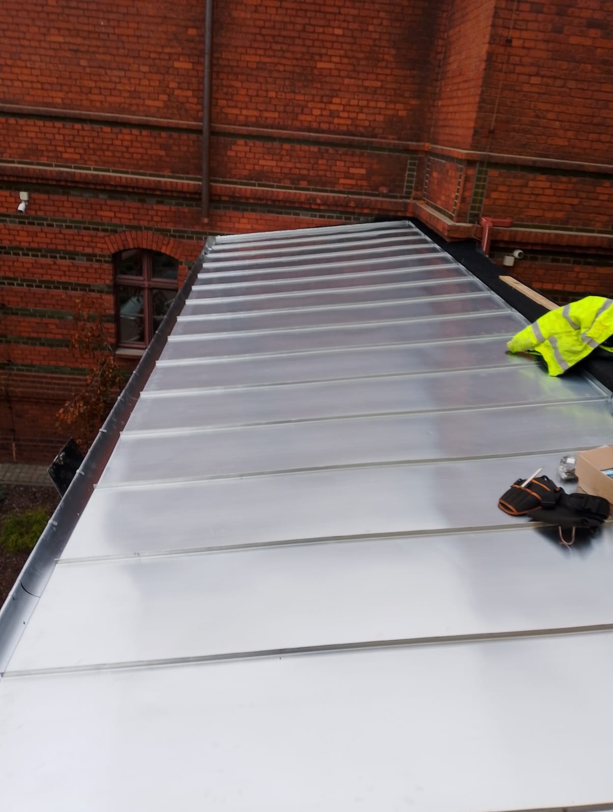 Nowe, błyszczące, metalowe pokrycie dachu z widoczną ceglaną ścianą budynku w tle. Na dachu leżą rękawice robocze i żółta kamizelka odblaskowa.