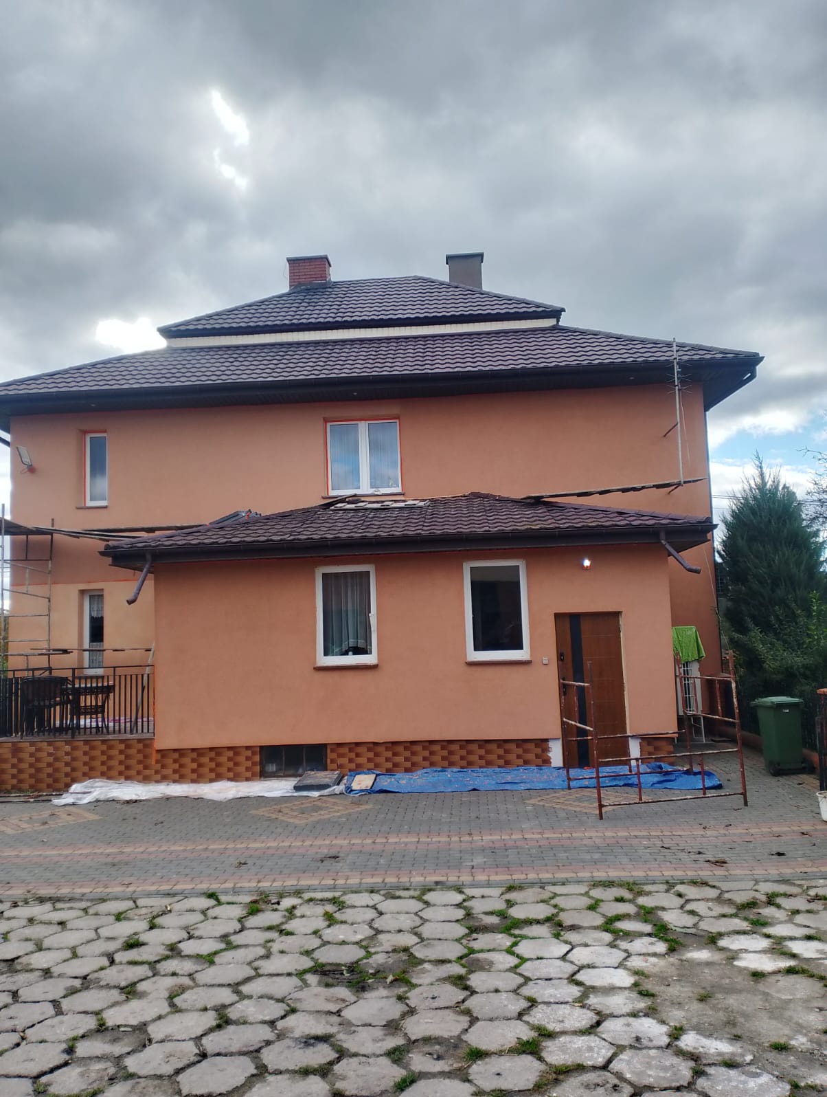 Dwupiętrowy dom z brązowym dachem i pomarańczową elewacją, widoczny z zewnątrz, z elementami rusztowania i zabezpieczeń na posesji. Pochmurne niebo w tle.