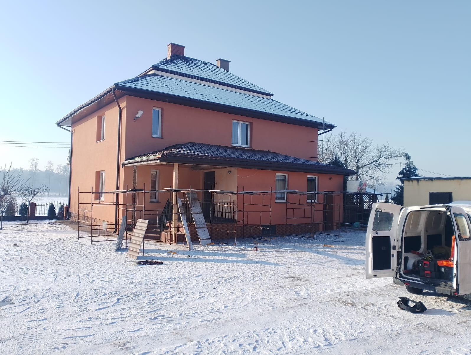 Ocieplony dom z rusztowaniem w zimowej scenerii. Widoczny otwarty samochód dostawczy z narzędziami. Dach pokryty ciemną dachówką. Słoneczny dzień.