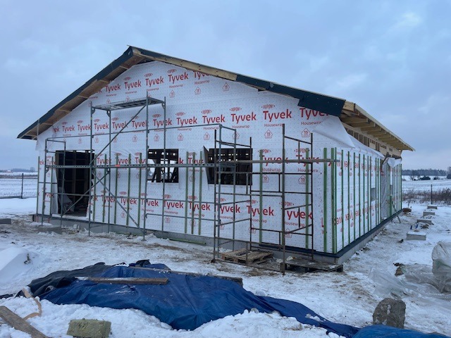 Dom w trakcie budowy, zabezpieczony folią Tyvek, z widocznymi rusztowaniami i elementami konstrukcyjnymi dachu. Zimowy krajobraz w tle.