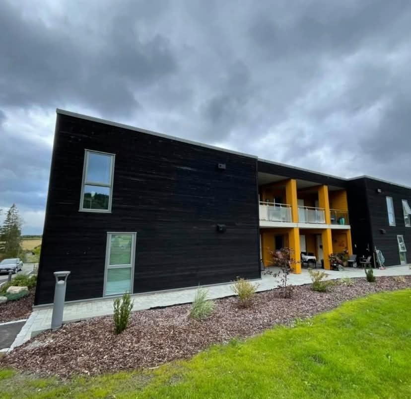 Dwukondygnacyjny budynek z ciemną, drewnianą elewacją i żółtymi balkonami, otoczony zielenią i korą drzewną, pod pochmurnym niebem.