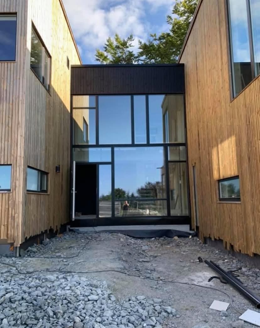 Nowoczesny budynek z drewnianą elewacją i dużymi przeszkleniami, widok z zewnątrz na teren w trakcie prac wykończeniowych.