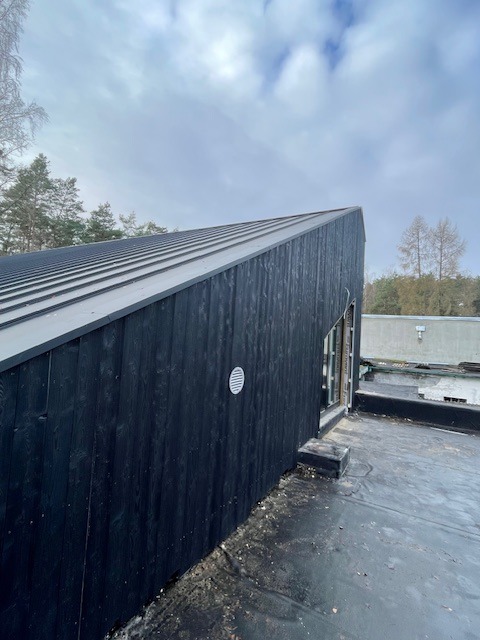 Nowoczesny dom z ciemną, drewnianą elewacją i metalowym dachem na tle pochmurnego nieba. Widoczny fragment dachu i ściany z wentylacją.