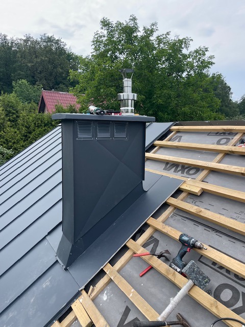 Szary komin z blachy na dachu w trakcie budowy, widoczne narzędzia i elementy konstrukcyjne. Komin z metalowym zakończeniem na tle zieleni.