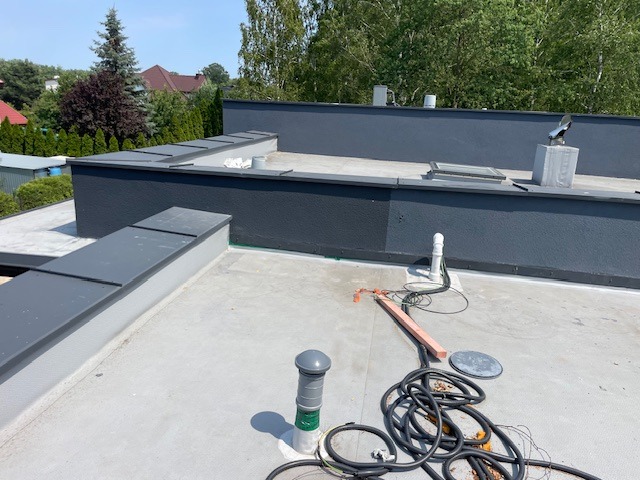 Szary, płaski dach z obróbkami blacharskimi i elementami instalacji. Widoczne detale wykończenia i zabezpieczenia dachu. Dach w otoczeniu zieleni.