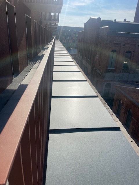 Długi, płaski dach z szarej blachy trapezowej, widziany z boku, z metalową balustradą w kolorze ceglastym. W tle budynki z cegły i błękitne niebo.