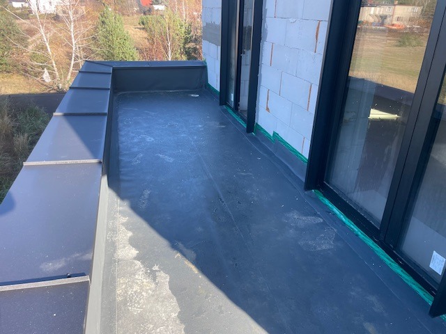 Wykończony, płaski dach z czarną membraną i obróbkami blacharskimi, widok z góry. Nowoczesne okna balkonowe z czarnymi ramami. Budynek w trakcie budowy.