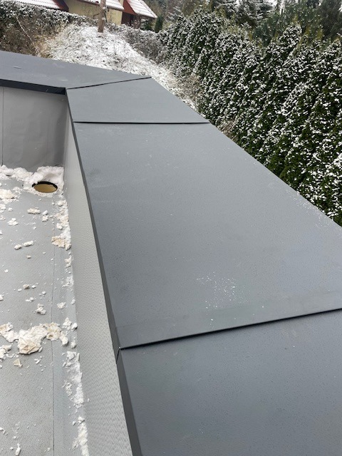 Nowoczesne, grafitowe obróbki blacharskie na dachu budynku, widoczne z bliska, z delikatnym oprószeniem śniegiem i zielenią w tle.