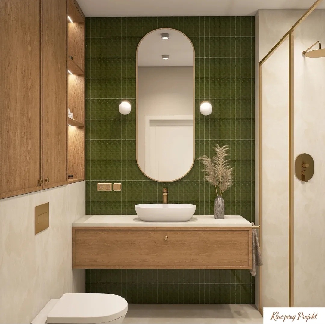 Nowoczesna łazienka z zielonymi płytkami, owalnym lustrem i drewnianą szafką pod umywalką. Minimalistyczny design z akcentami złota i subtelnym oświetleniem.