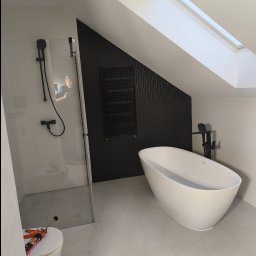 Cezary Toruński - Nowoczesna łazienka na poddaszu z wolnostojącą wanną, kabiną prysznicową typu walk-in i czarnym grzejnikiem drabinkowym na tle ściany z pionowymi panelami.