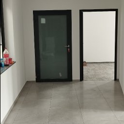 Cezary Toruński - Wnętrze z szarą, kwadratową podłogą z płytek, dwoje czarnych drzwi i białymi ścianami. Widoczne butelki na parapecie. Sufit kasetonowy.