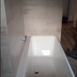 Cezary Toruński - Biała wanna z niebieską taśmą zabezpieczającą krawędzie, zamontowana w surowej łazience z szarymi płytkami imitującymi beton. Widoczne elementy instalacji.