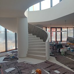 Cezary Toruński - Wnętrze w trakcie malowania: spiralne schody z drewnianą balustradą, zabezpieczone podłogi, okna z folią ochronną. Widoczne puszki z farbą i narzędzia malarskie.