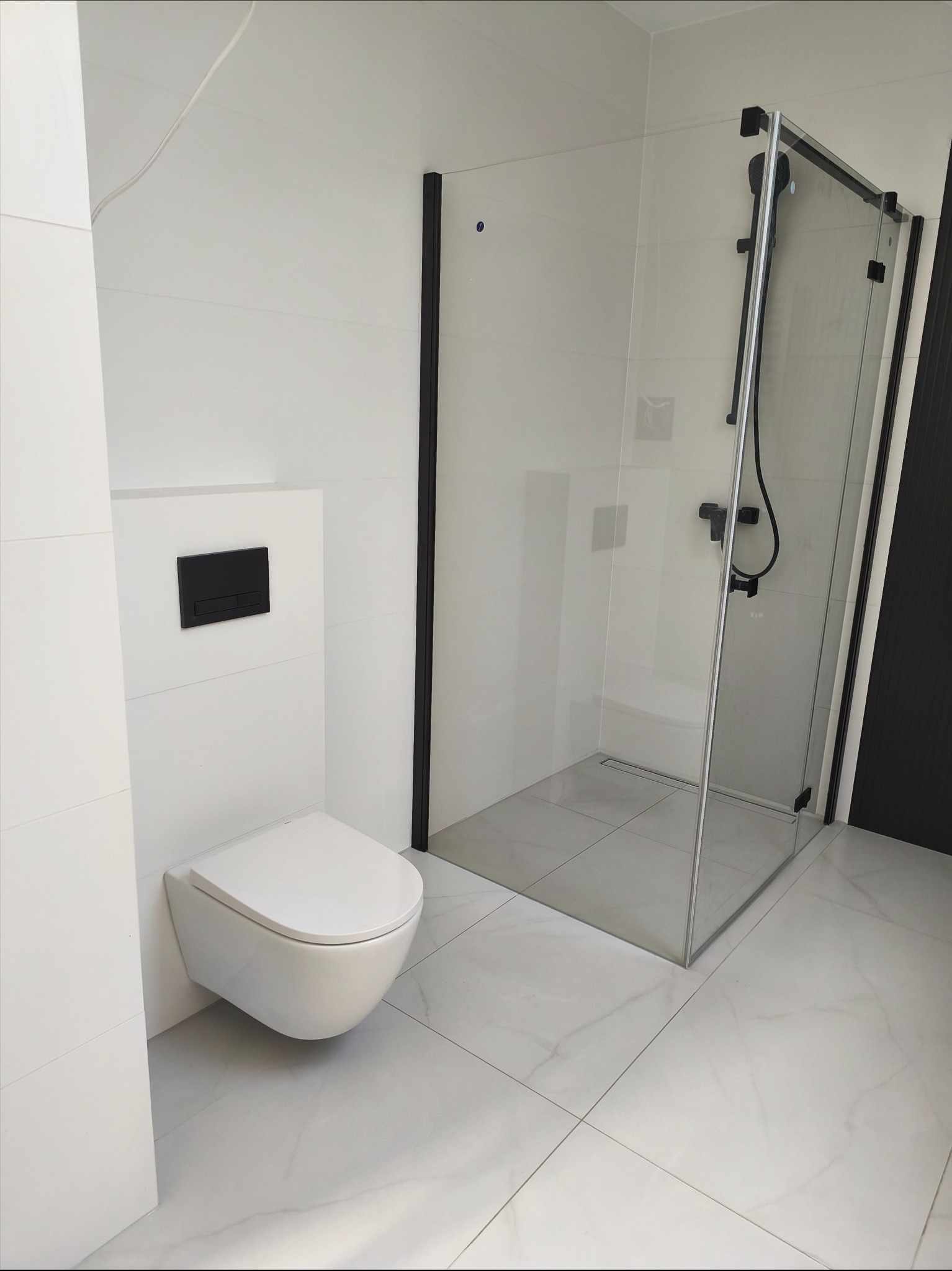 Nowoczesna łazienka z podwieszaną toaletą i kabiną prysznicową typu walk-in z czarnymi profilami. Białe płytki imitujące marmur na podłodze i ścianach.