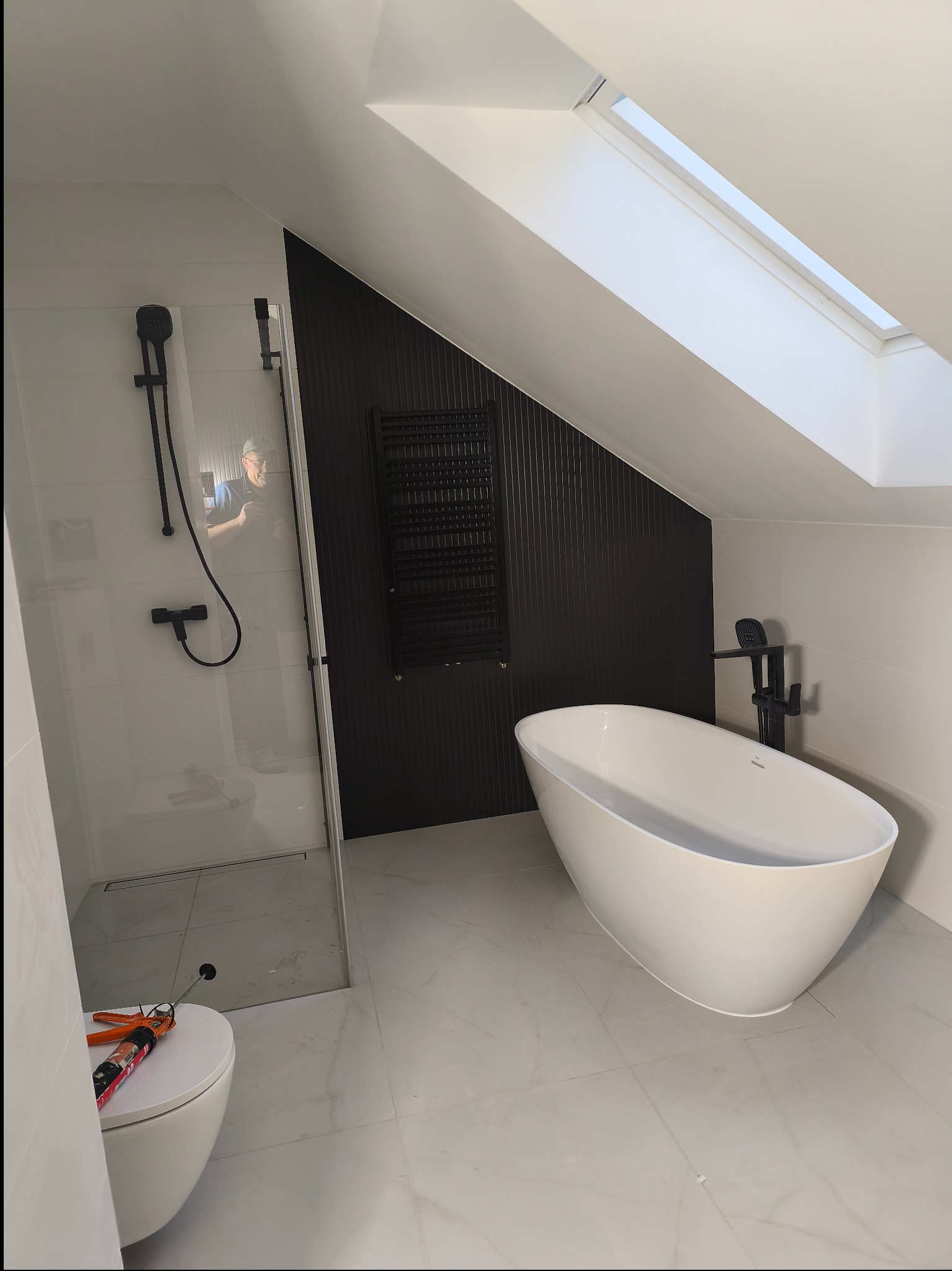 Nowoczesna łazienka na poddaszu z wolnostojącą wanną, kabiną prysznicową typu walk-in i czarnym grzejnikiem drabinkowym na tle ściany z pionowymi panelami.