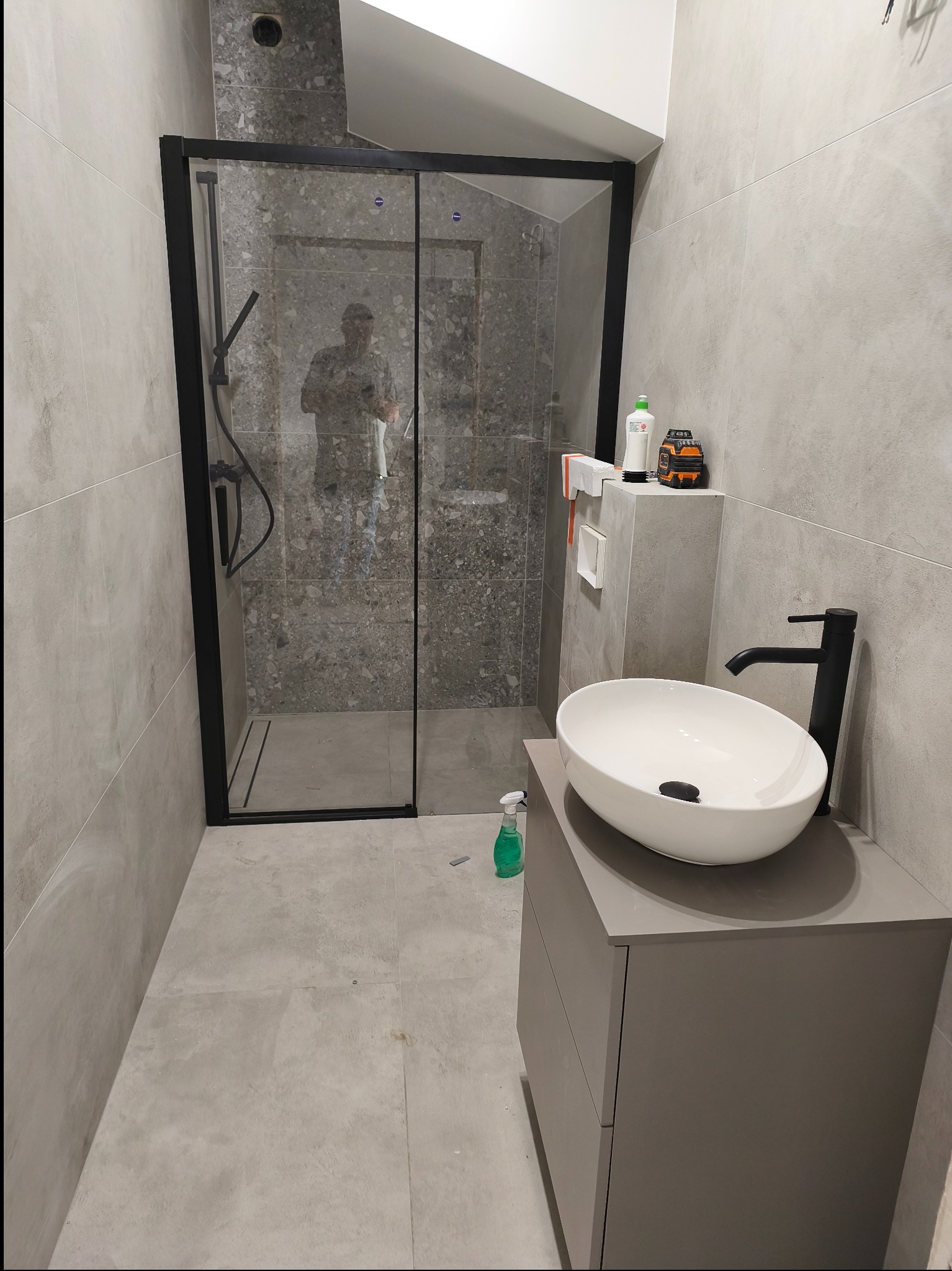 Nowoczesna łazienka z kabiną prysznicową typu walk-in z czarną ramą, umywalką nablatową na szarej szafce i szarą glazurą na ścianach. Widoczne akcesoria łazienkowe.